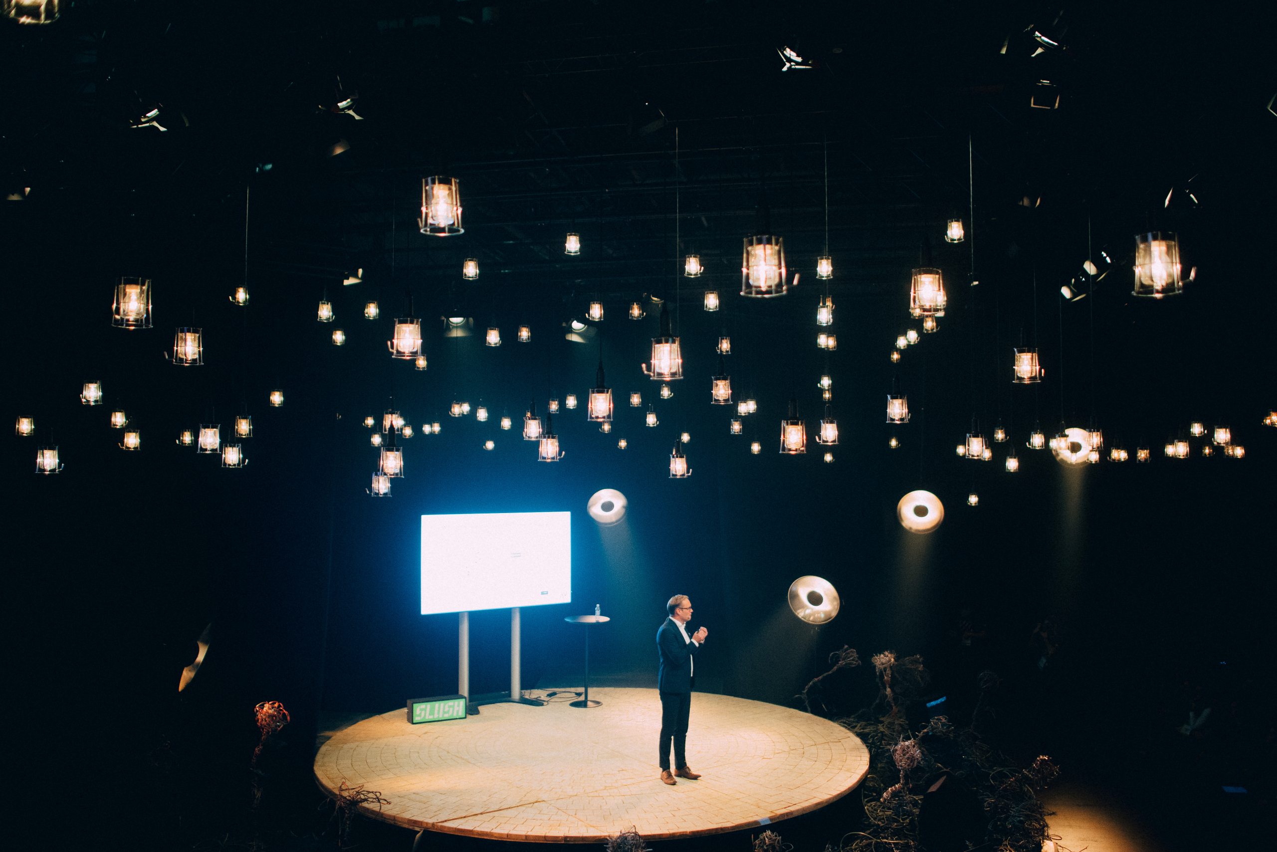 Single speaker on a small round Slush stage beneath a canopy of hanging lantern lights in a dark hall