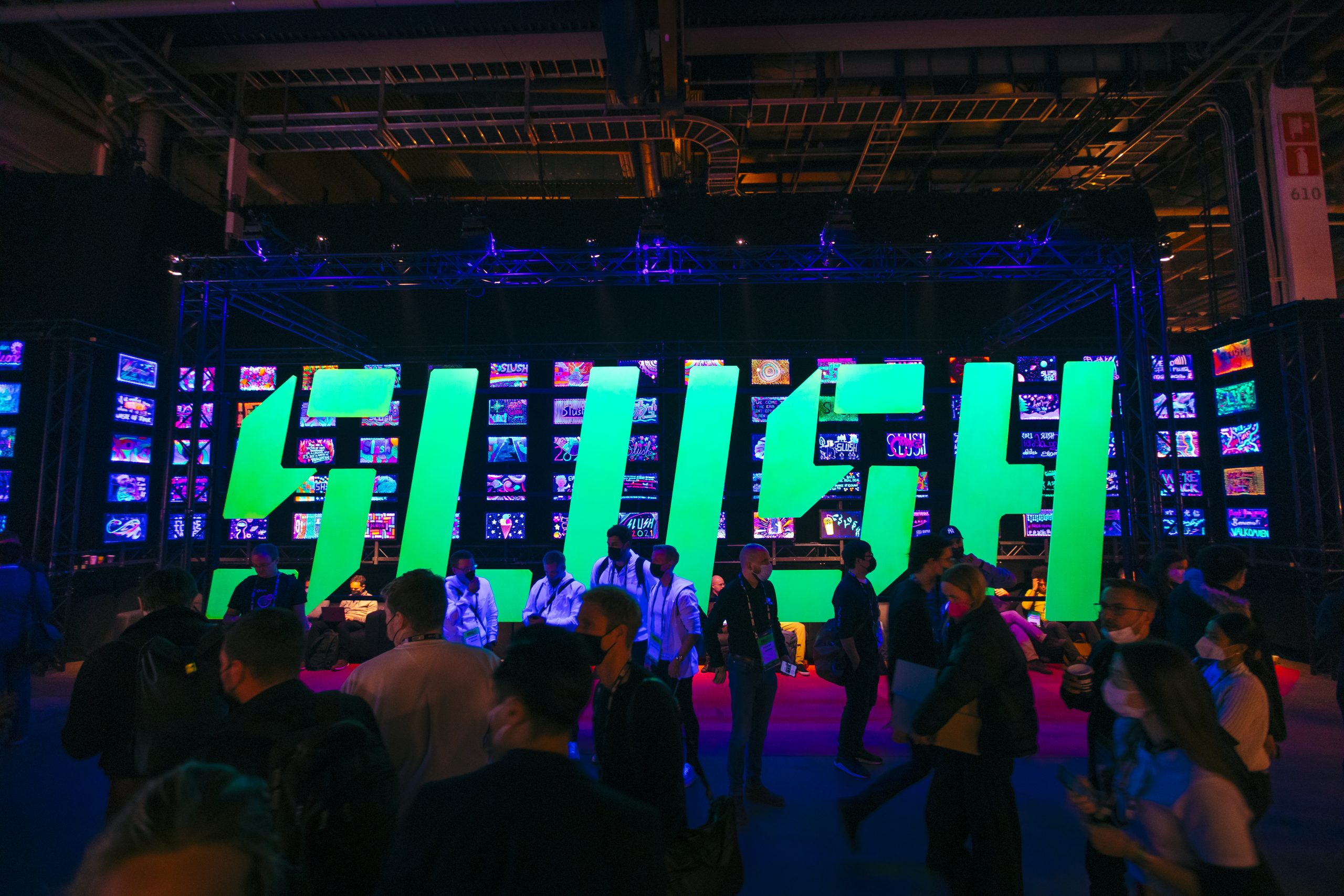 Illuminated green ‘SLUSH’ sign built from stacked screens, with attendees mingling in front