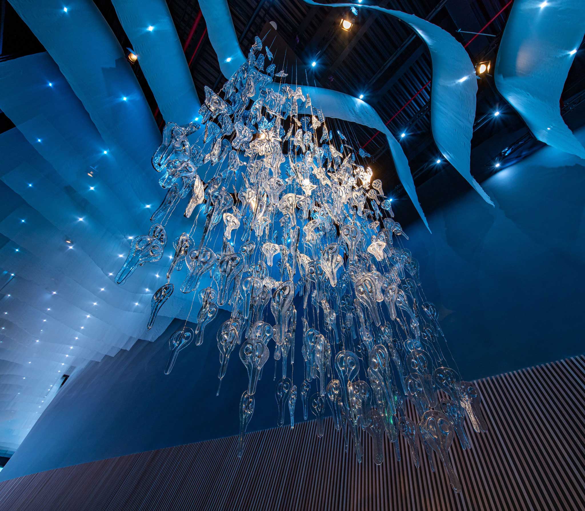 Cascading glass chandelier installation suspended in the blue-lit angular interior of the Finland pavilion