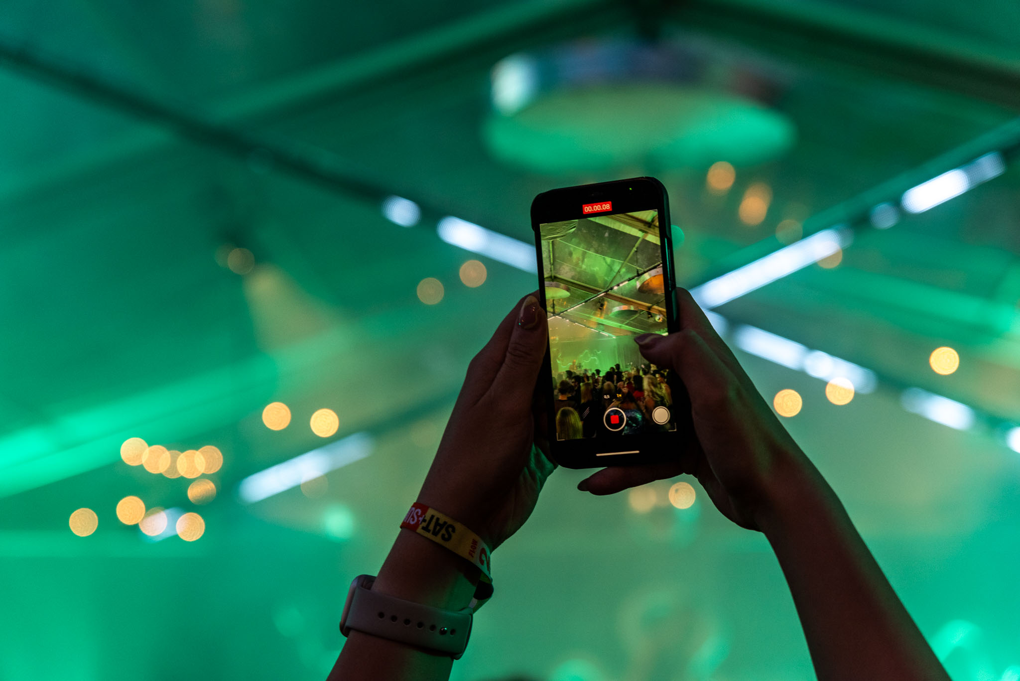 Festival-goer filming on a phone under green beam lighting and warm bokeh lights at Flow Festival