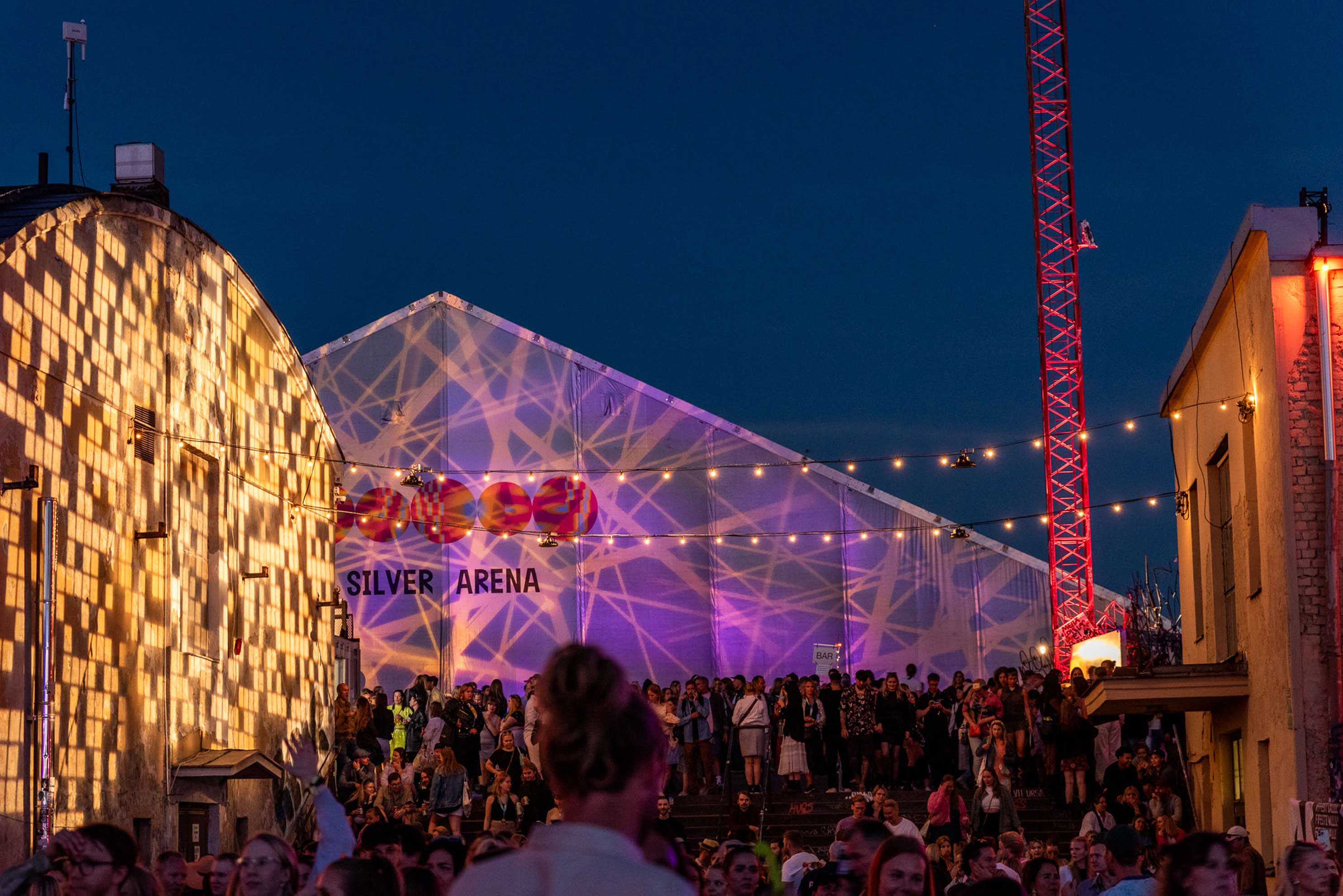 Crowd outside Flow Festival’s tent stage at dusk, lit pink and strung with warm festoon lights beside the industrial buildings