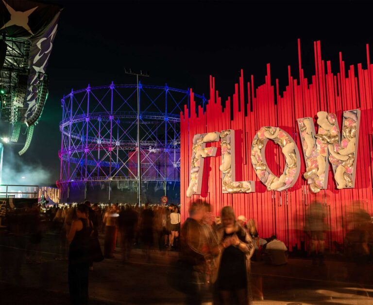 Flow Festival’s ‘FLOW’ lettering against red strips beside the blue-lit gasometer at night