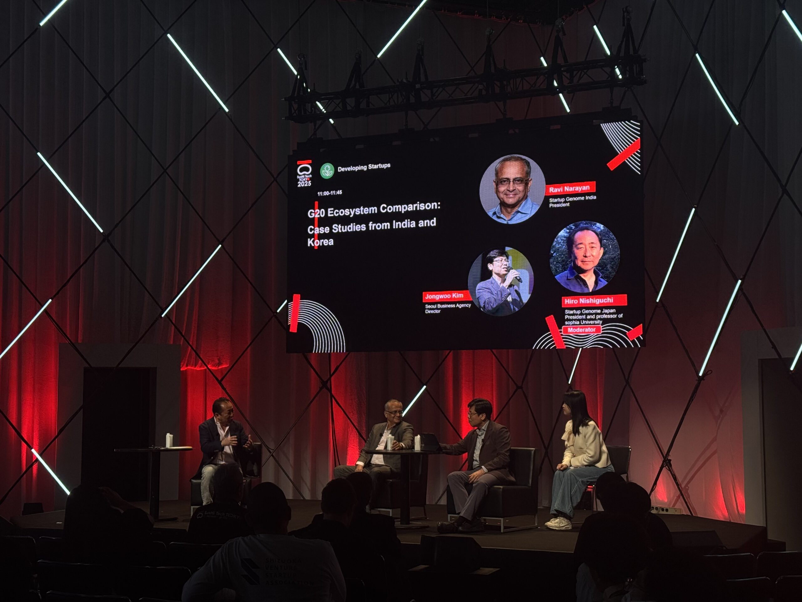 Panel session at SusHi Tech with speakers on stage below a large screen of session details and red diagonal lighting on the walls