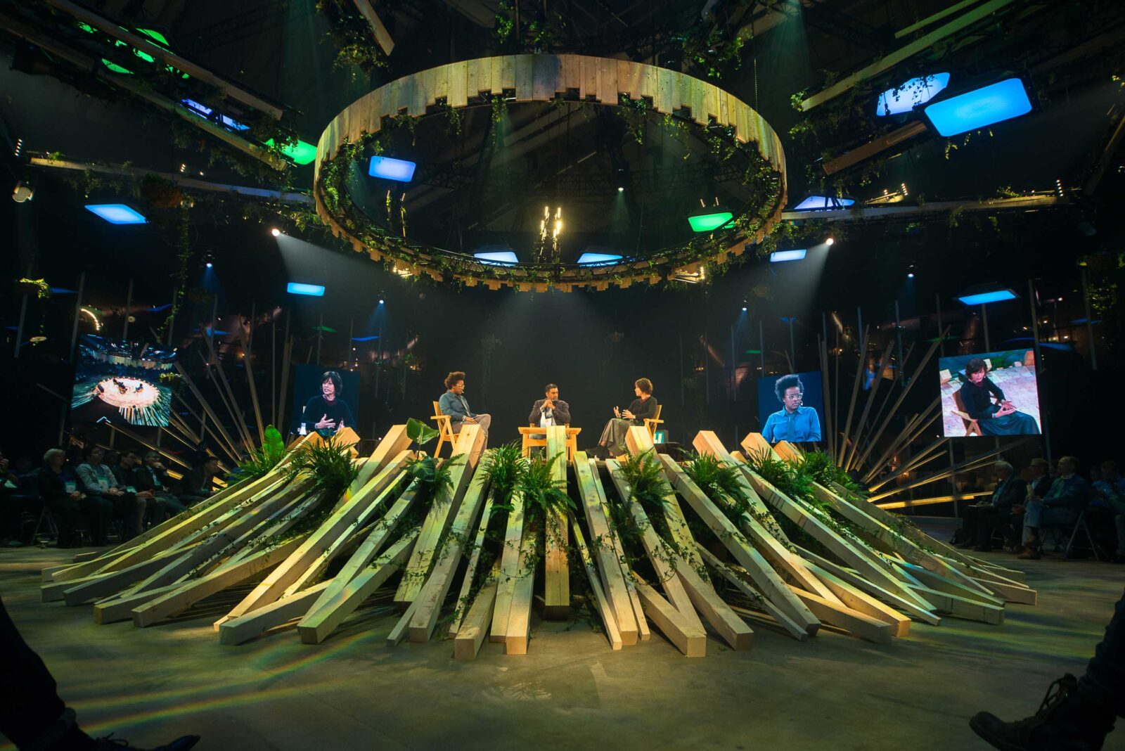 Event stage under a circular halo light with speakers seated among bundles of leeks and plants radiating across the stage