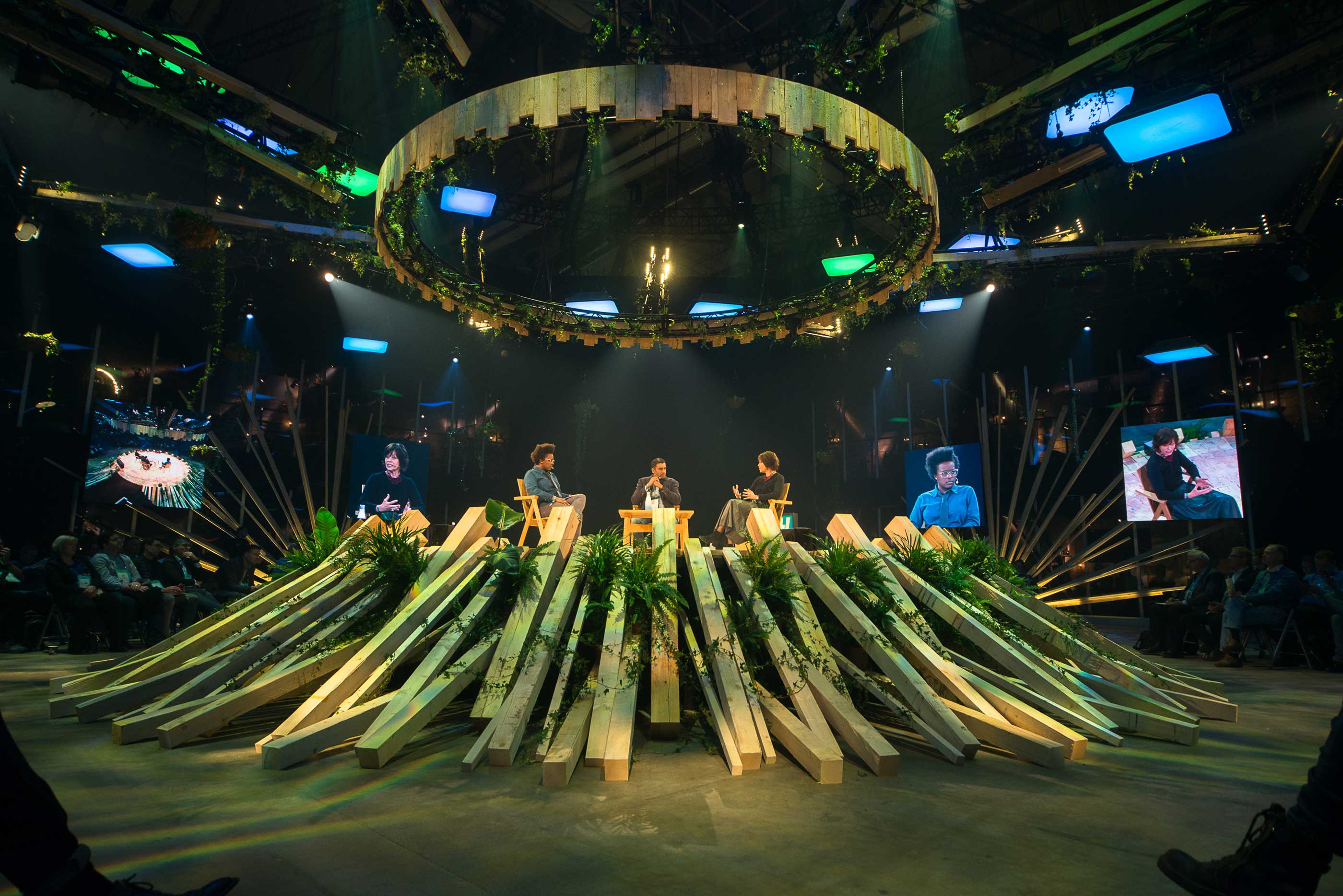 Slush stage under a large circular halo light, with speakers seated among bundles of leeks and plants radiating across the stage
