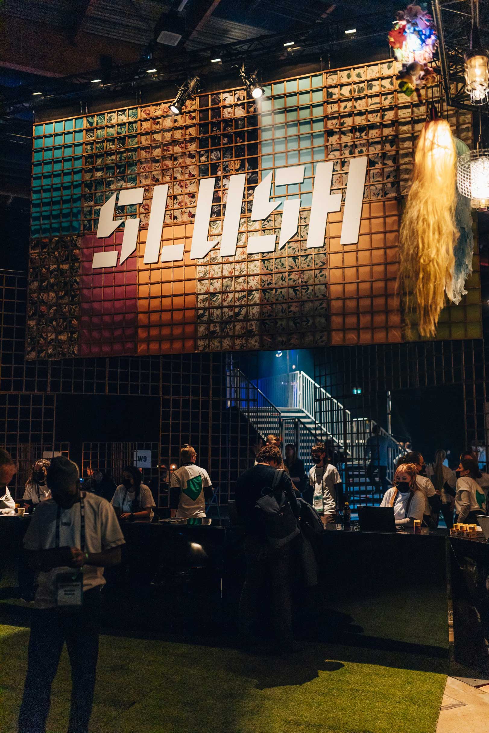 Large ‘SLUSH’ sign on a textured tiled wall above an industrial staircase, with attendees mingling below