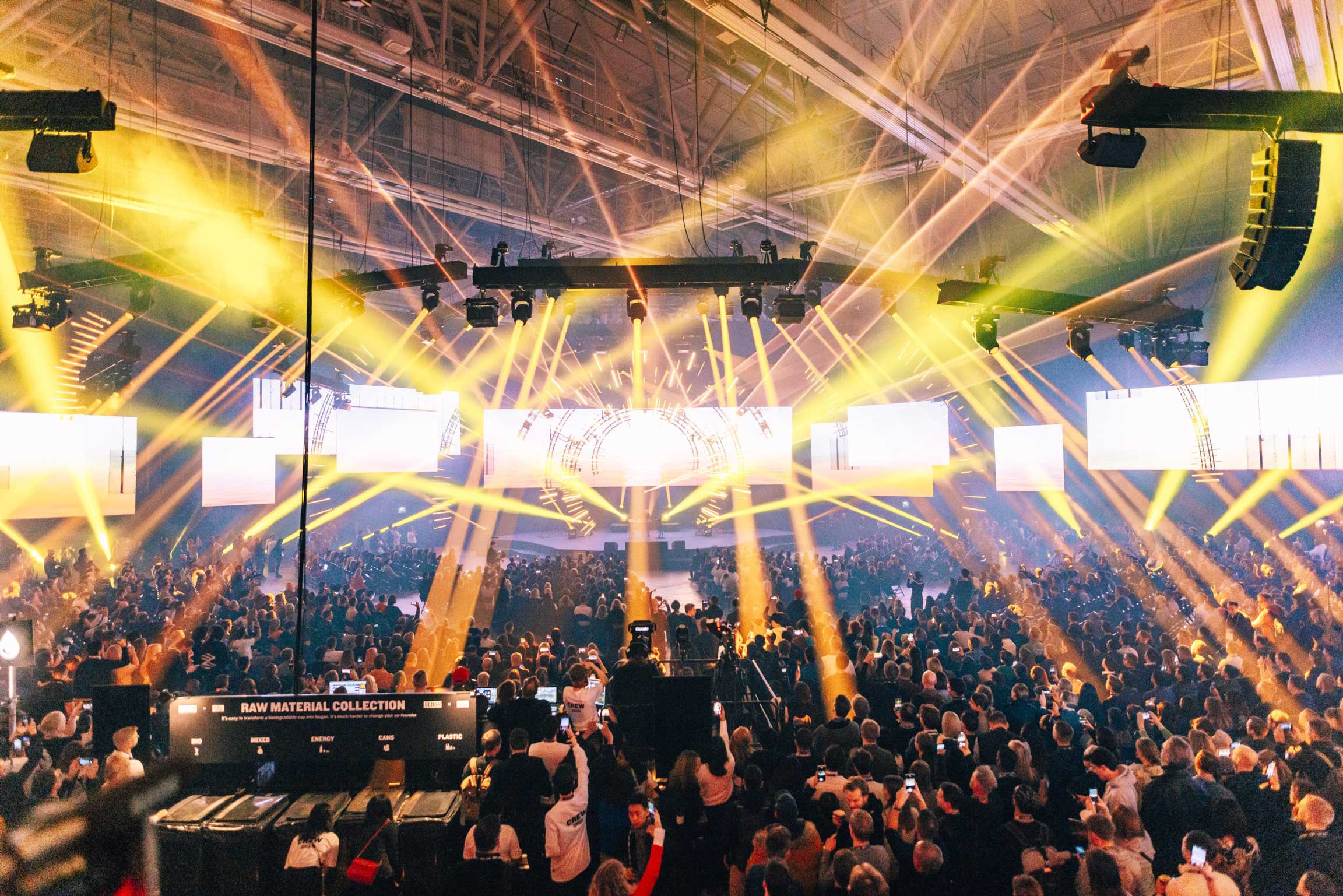 Slush stage and crowd bathed in radiating golden beam lighting across the hall