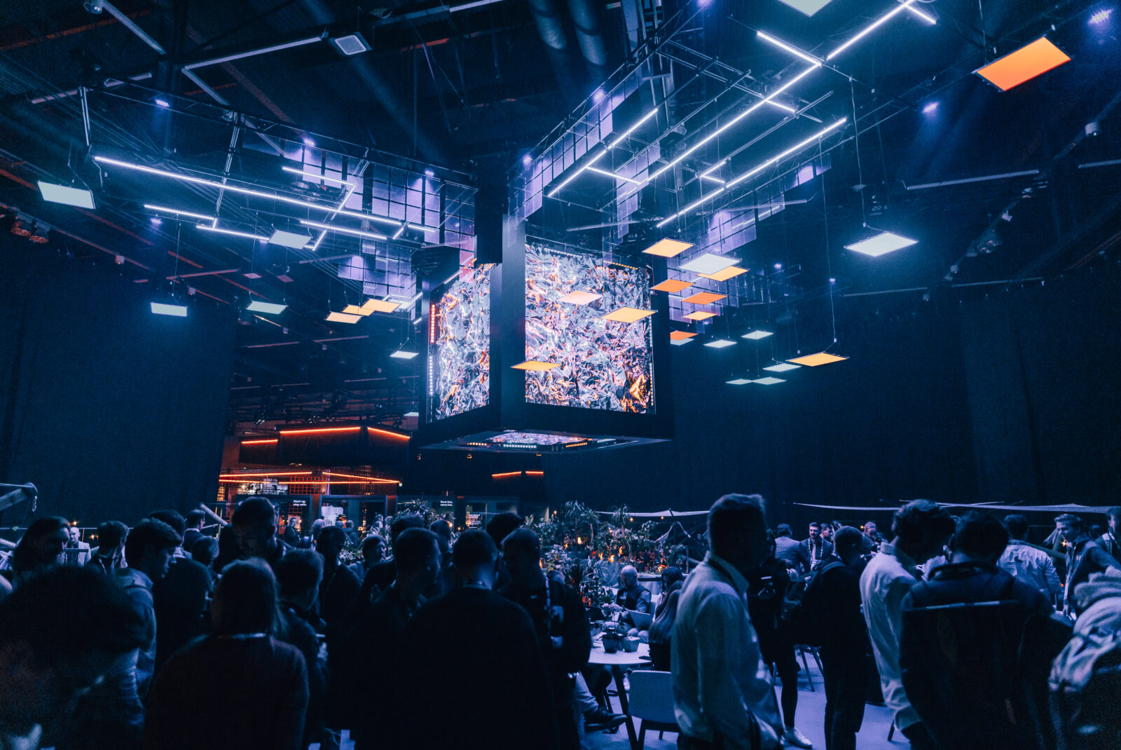 Event hall with a large suspended screen and mixed lighting above a mingling crowd