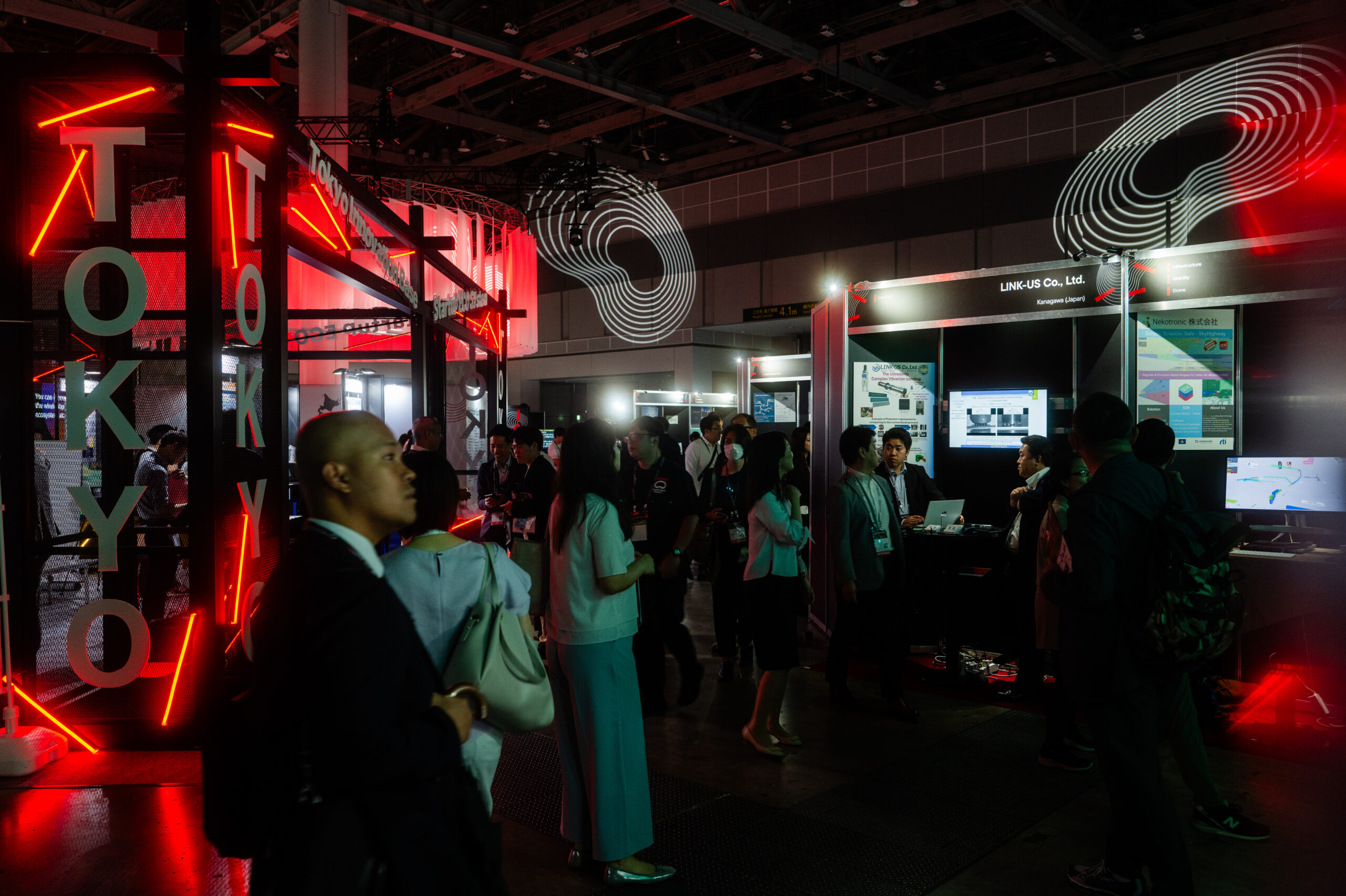 Dark exhibition hall at SusHi Tech with red neon accents, a vertical ‘TOKYO’ sign and spiral graphics as attendees explore the stands