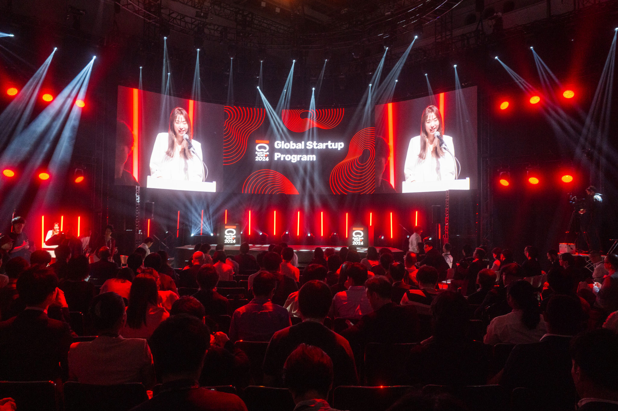 SusHi Tech main stage under sweeping beam lights, two speakers shown on a large ‘Global Startup Program’ screen above a full audience