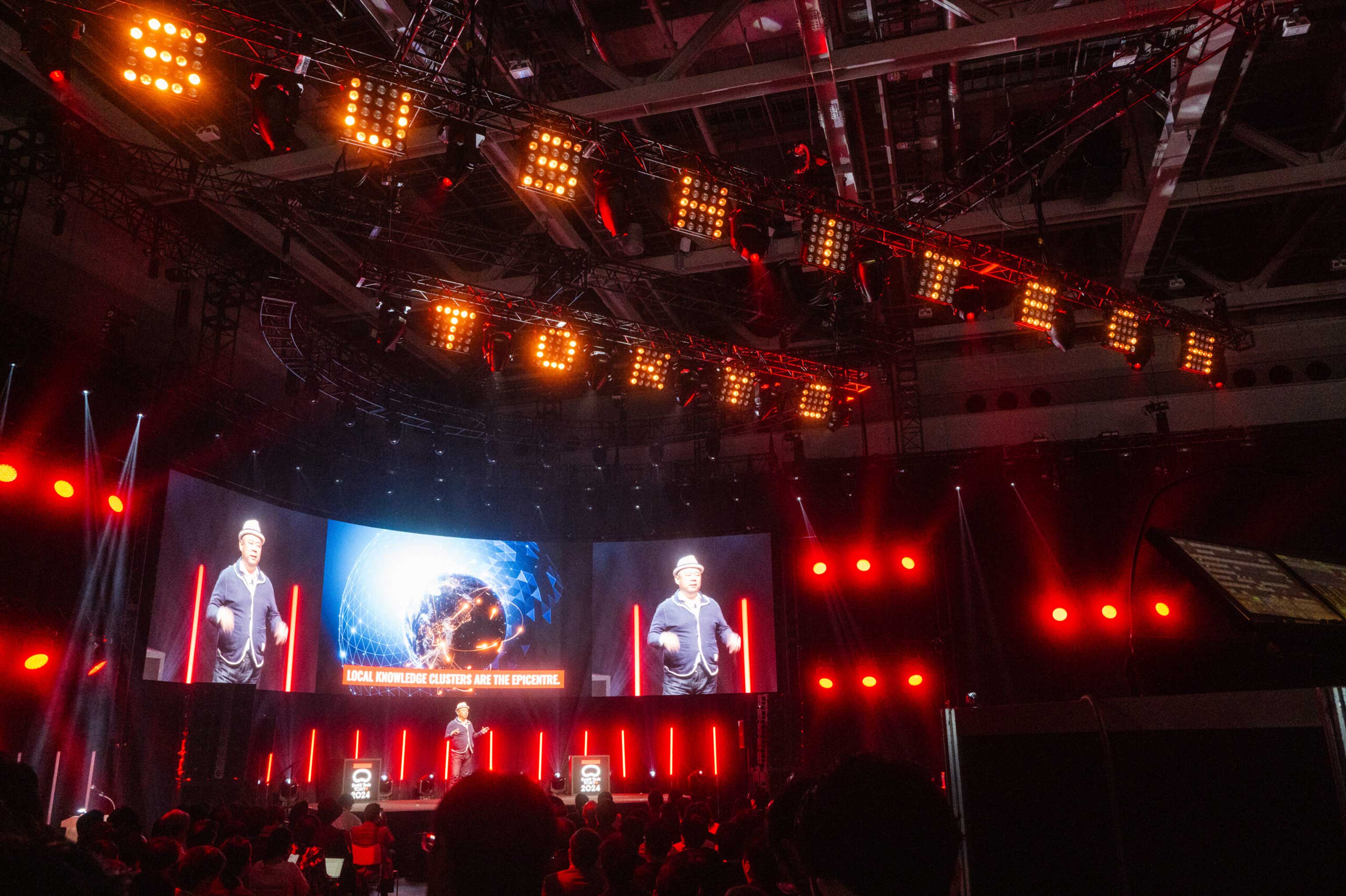 Wide view of the SusHi Tech main stage under warm orange lighting, with a speaker, large screens and an overhead lighting rig