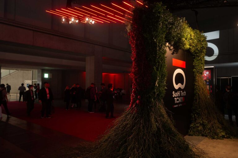 Column wrapped in greenery beside the SusHi Tech logo under red lighting, with attendees mingling