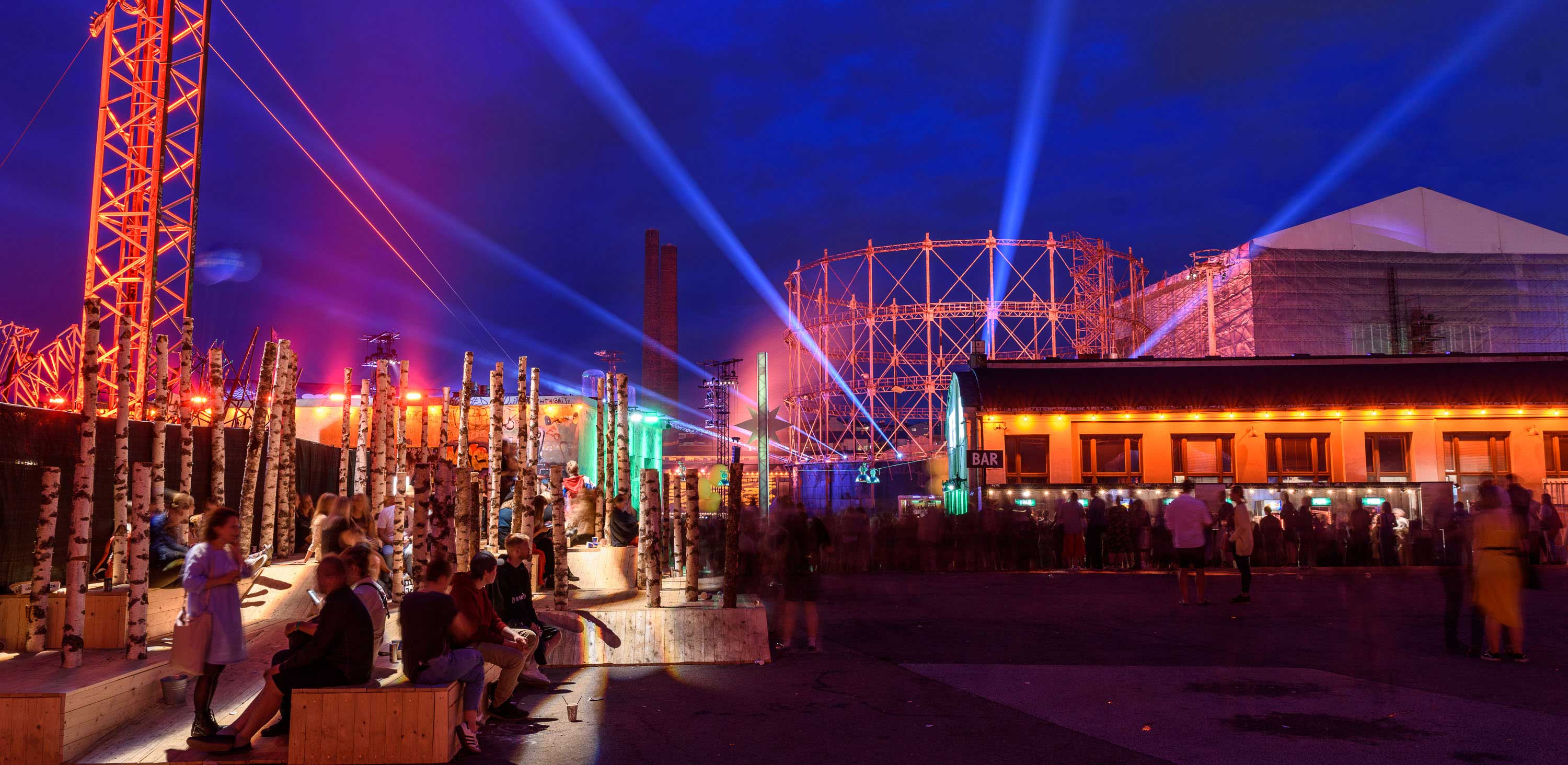 Blue laser beams over the industrial gasometer frame at night at Flow Festival, with warm-lit bars and a crowd below