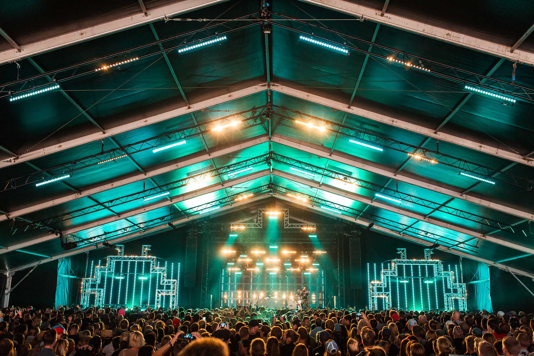 Interior of Flow Festival’s large tent stage lit teal along the roof beams, with a lighting rig above a dense crowd