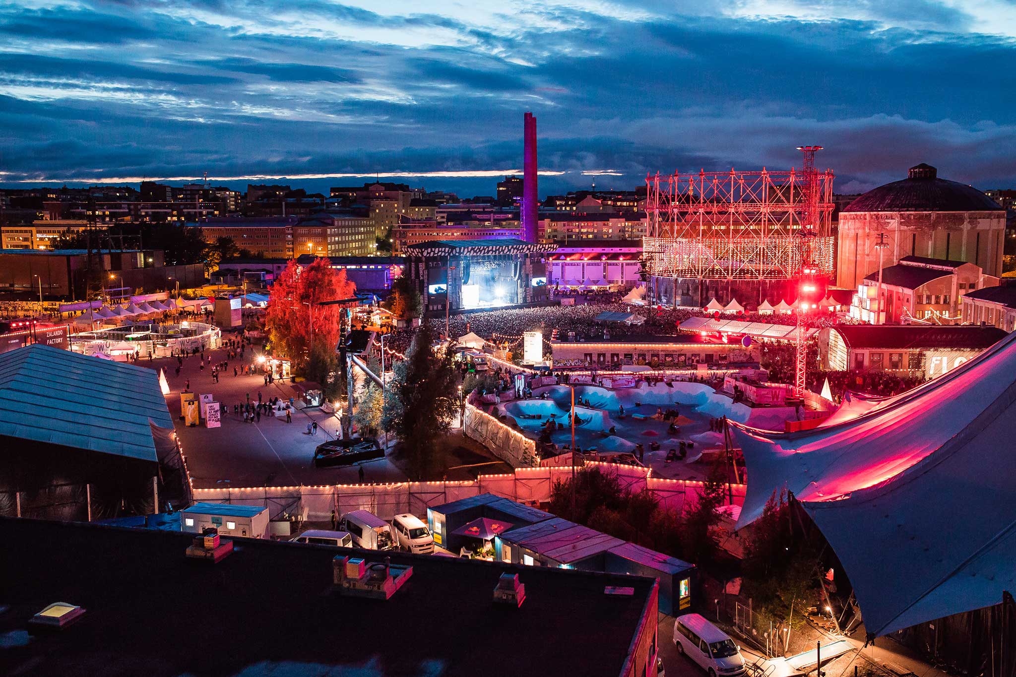 Elevated dusk view of the Flow Festival grounds on the former Suvilahti power-plant site in Helsinki, with stages and colourful lighting across the area