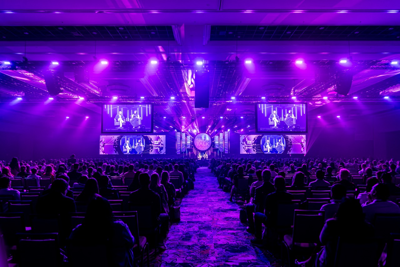 Shoptalk conference hall lit purple, with a central stage, large screens and a seated audience along the aisle