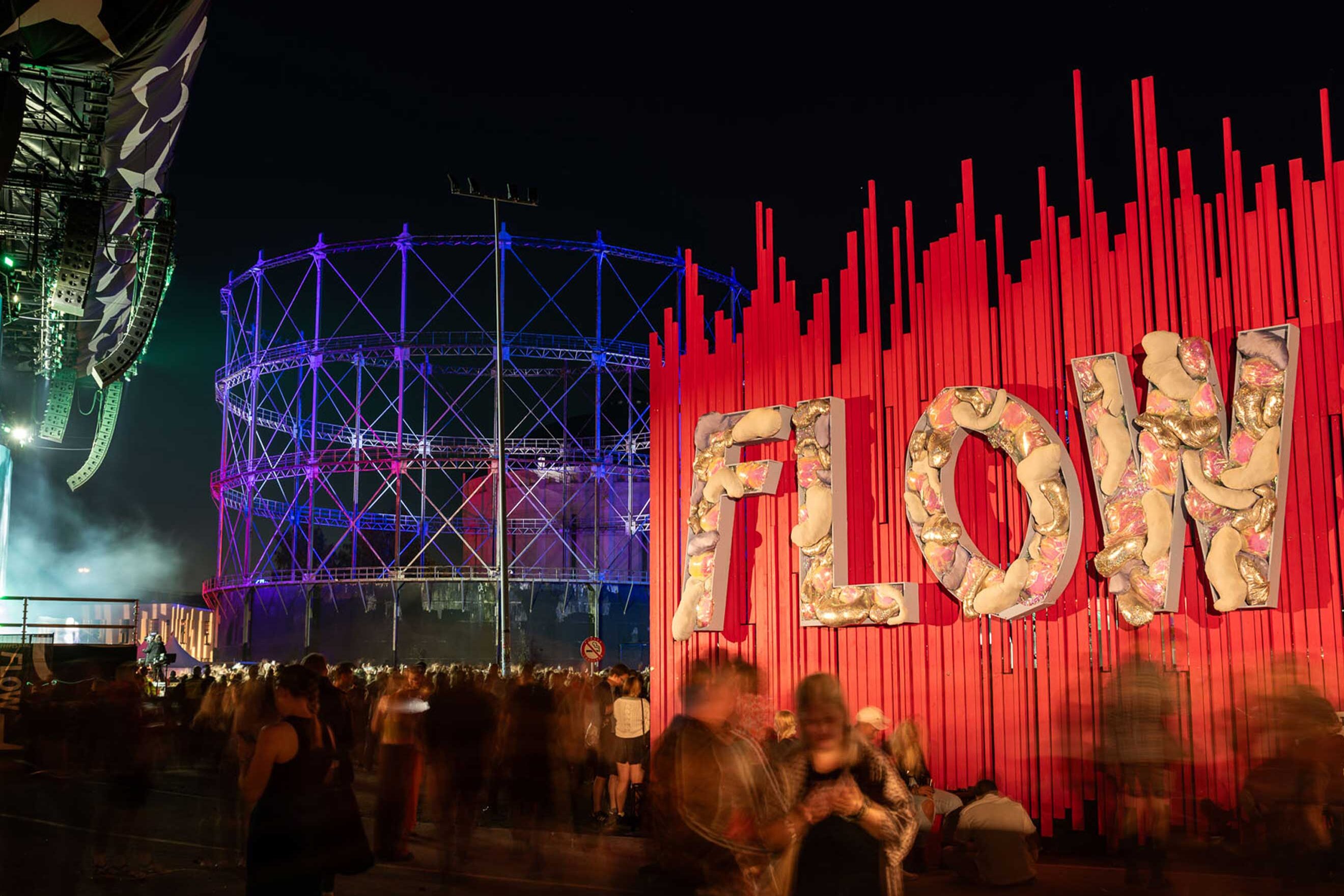 Flow Festival’s ‘FLOW’ lettering installation against red vertical strips beside the blue-lit gasometer at night