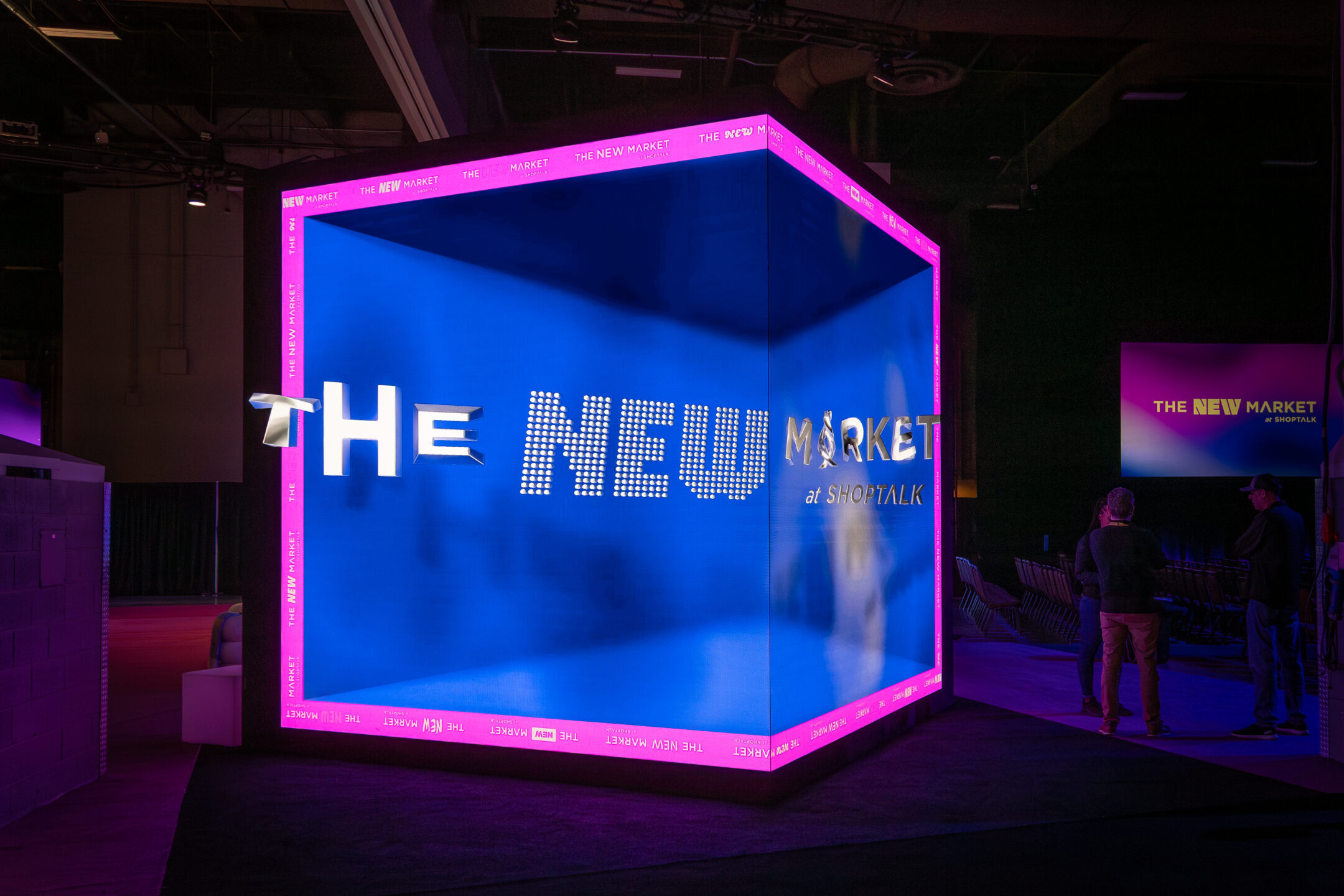Large blue illuminated cube with pink neon edges reading ‘THE NEW MARKET’ in the Shoptalk expo hall
