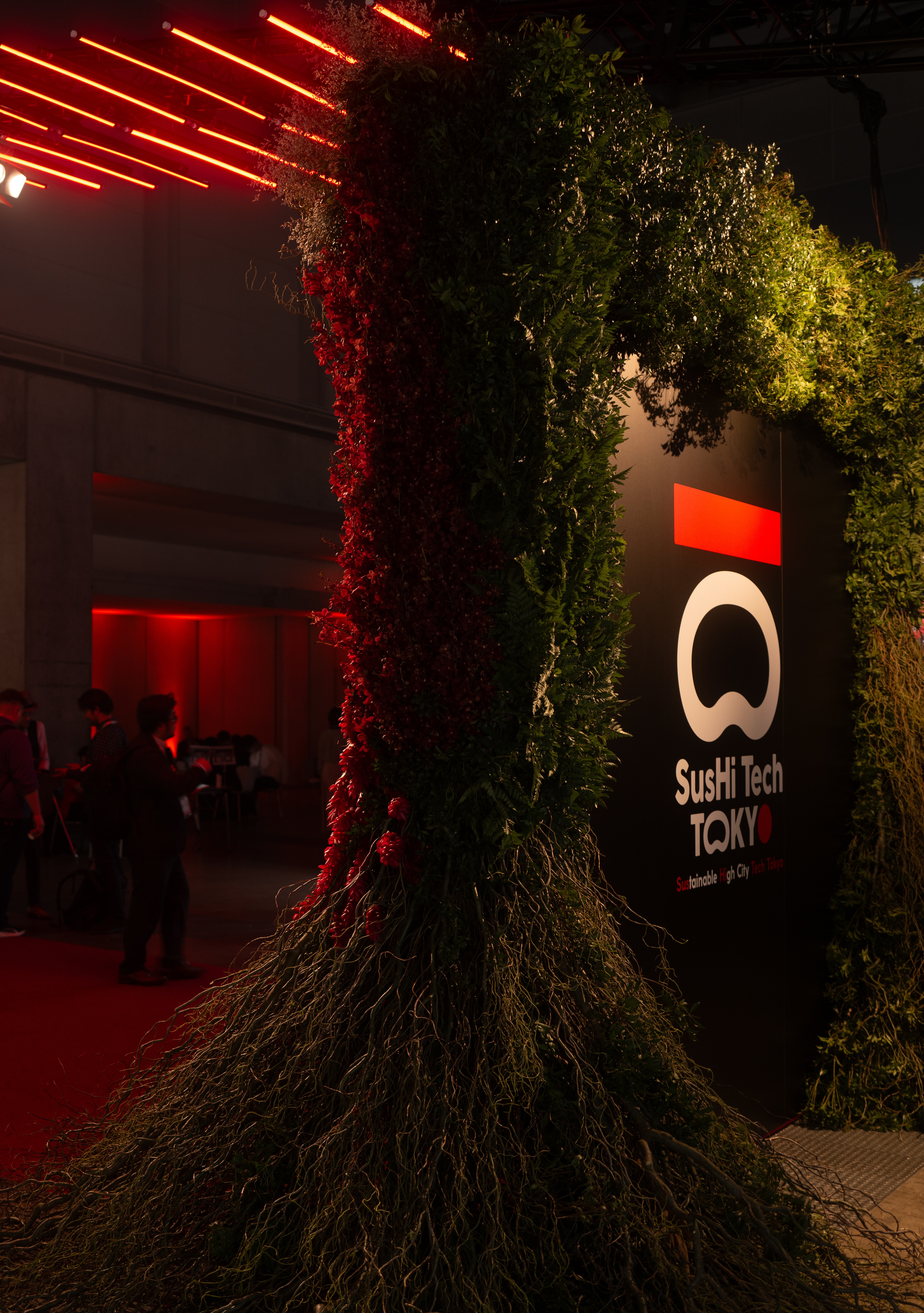 Column wrapped in greenery beside the SusHi Tech logo under red lighting, with attendees mingling