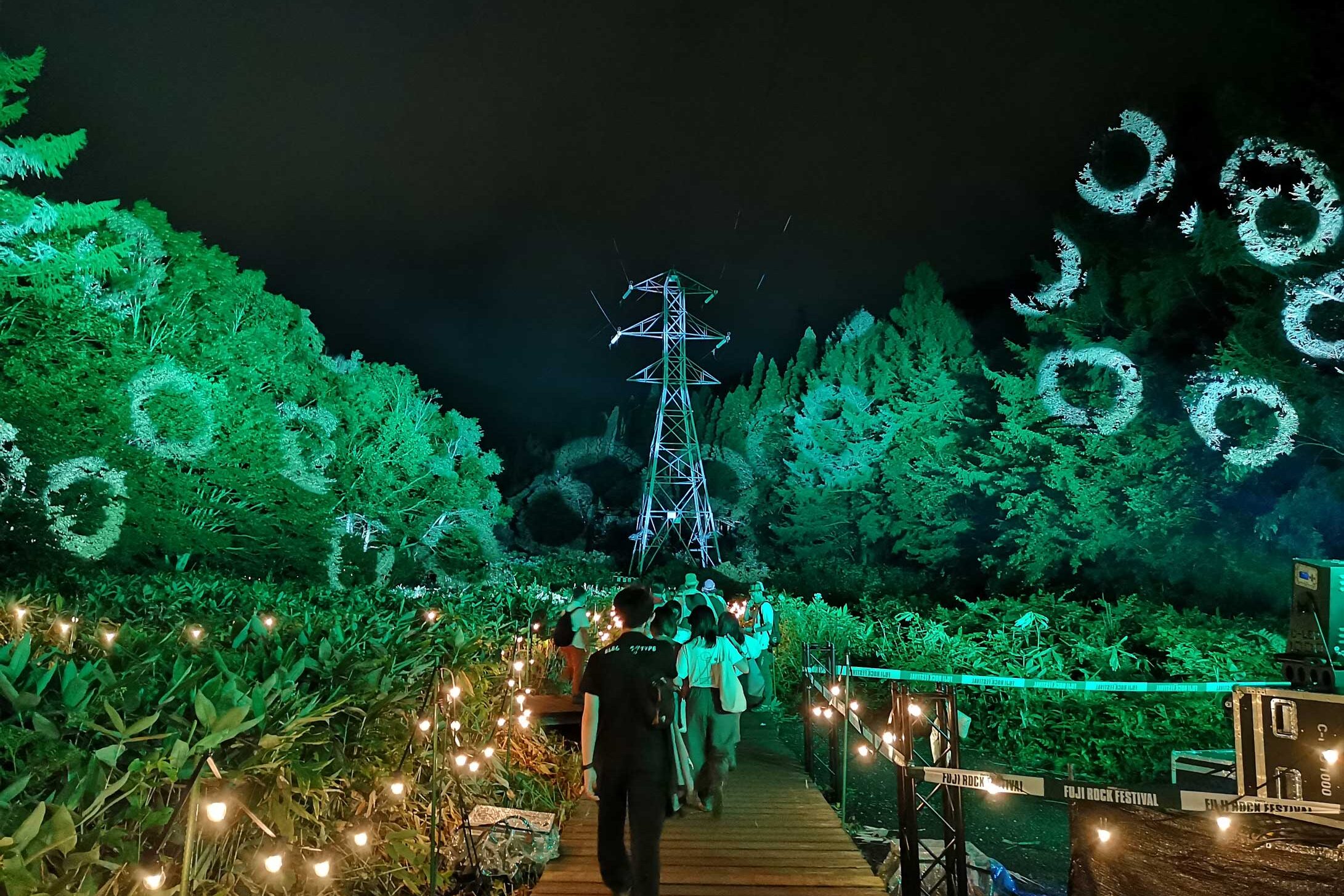 Night forest path at Fuji Rock beneath a transmission tower, with green lighting, hanging bulbs and glowing ring projections in the trees